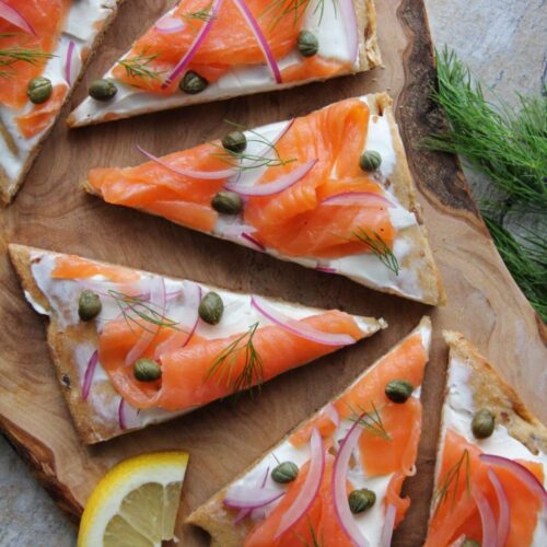 overhead view of smoked salmon flatbread pizza on a wood cutting board. Flatbread consists of cream cheese, slices of smoke salmon, capers, red onions and fresh dill.