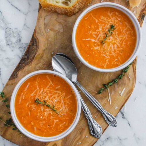 Overhead view of two bowls of creamy tomato soup. Bowls are resting on a wood cutting board and are surrounded by spoons, crusty bread and fresh thyme.