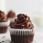 Close up view of a dark chocolate cupcake with a white paper liner. Other cupcakes seen in the background.