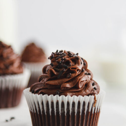 Close up view of a dark chocolate cupcake with a white paper liner. Other cupcakes seen in the background.