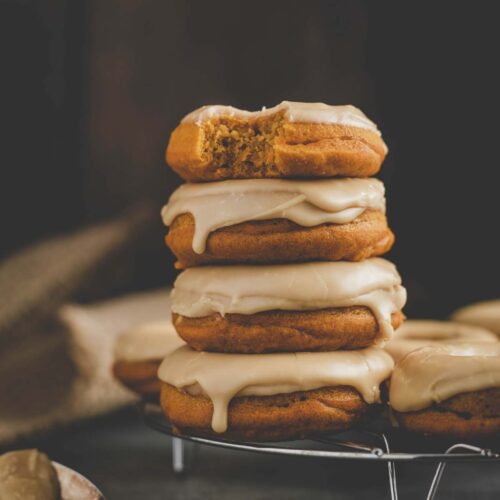 stack of maple glazed pumpkin donuts sitting on a wire rack with the top donut having a bite taken from it.