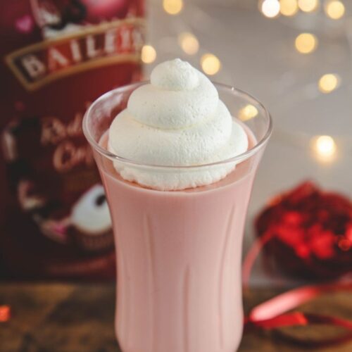 A champagne flute filled with baileys red velvet cupcake mousse, topped with whipped cream. Behind the cup is a bottle of Baileys and twinkling christmas lights.