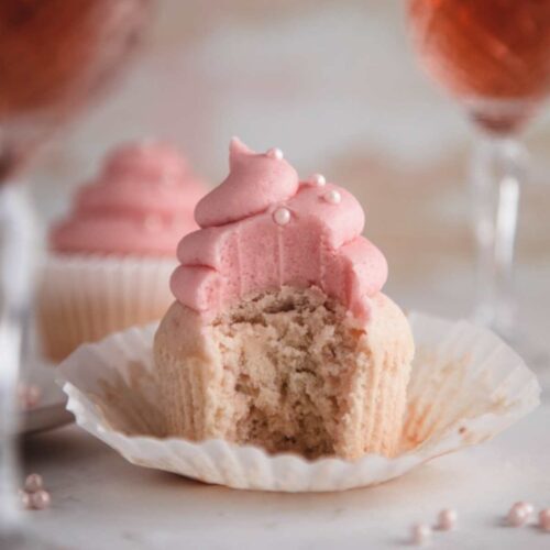 A rose wine cupcake placed on a paper cupcake liner. A large bite is taken from the front of the cupcake. Behind the cupcake are two tall glasses of rose wine.