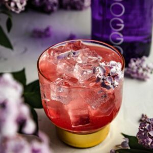 Glass of strawberry lilac gin, garnished with lilac flowers. Glass is surrounded with lilac flowers and a bottle of Indoggo gin.