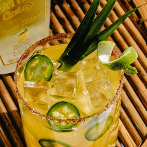 Close up view of a glass of spicy mango margarita. The cocktail is garnished with jalapenos, lime rind and tropical leaves.