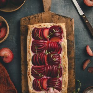 Overhead view of a puff pastry plum tart, resting on a wood cutting board. The board is surrounded with sliced plums, fresh thyme, a dark red linen cloth and knife. At the bottom of the page, a hand is placing a fresh sprig of thyme onto the tart.