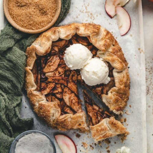 Overhead view of an apple galette with puff pastry. The galette is topped with two scoops of vanilla ice cream. The galette is surrounded by a bowl of raw sugar, apple slices, green linen and a cup of flour.