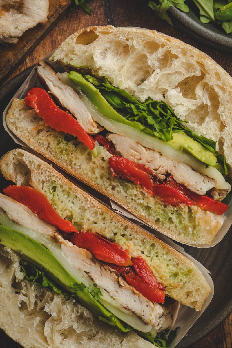 Overhead image of sliced tuscan turkey sandwich, surrounded by various ingredients including grilled turkey breast, roasted red peppers, baby arugula, ciabatta bun.