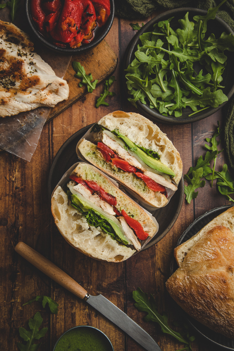 Overhead image of sliced tuscan turkey sandwich, surrounded by various ingredients including grilled turkey breast, roasted red peppers, baby arugula, ciabatta bun.
