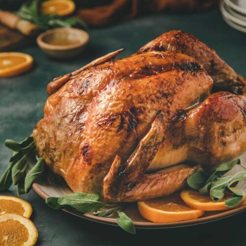 full roasted bourbon glazed turkey in a large serving plate. The turkey is surrounded by sage leaves, orange clised and plates.