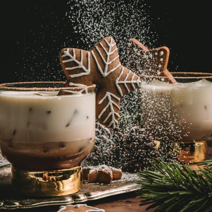 two glasses of ginerbread white russians garnished with star shaped gingerbread cookies. Icing sugar is being sprinkles from above.