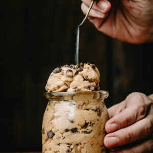 A hand holding a spoon, scooping edible chocolate chip cookie dough out of a clear jar. Jar is placed on a wood surface, surrounded by mini chocolate chips.