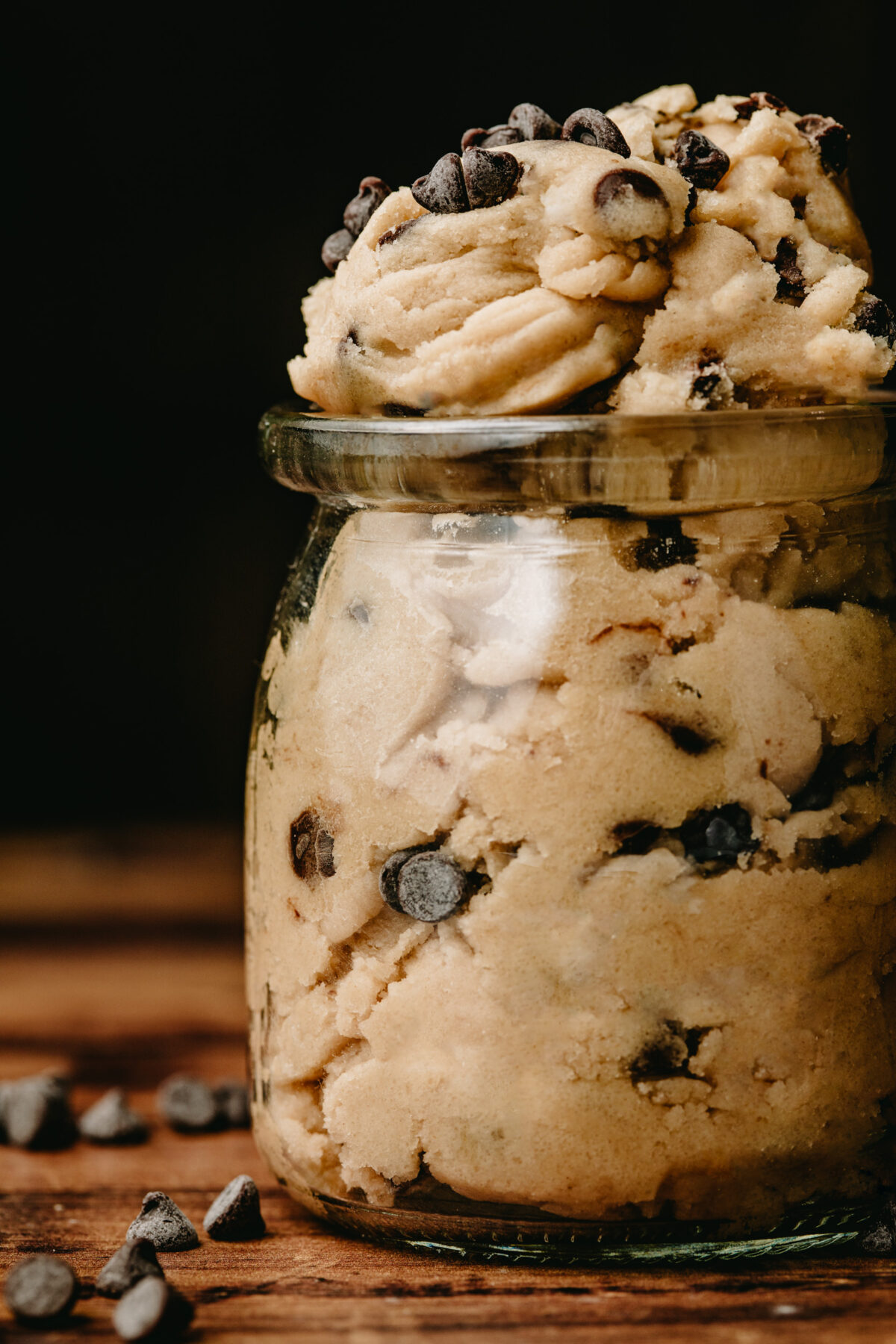 Close up view of jar filled with edible chocolate chip cookie dough on a wood surface, with mini chocolate chips scattered around it.