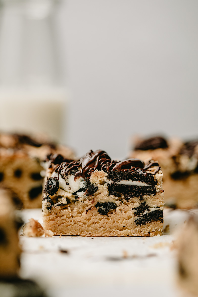 Front view of a piece of oreo blondie, surrounded by other pieces. In the background, a clear bottle of milk is seen in the left corner.