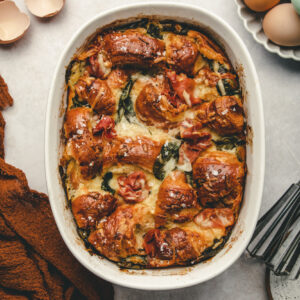 Overhead view of Prosciutto and Spinach Strata dish, surrounded with eggs and linen.