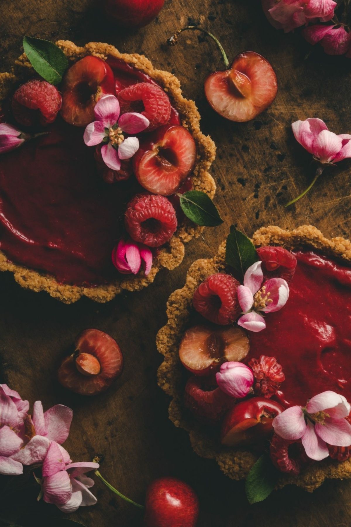 Overhead view of two mini raspberry cherry tarts on a wood board, surrounded by pink flowers and sliced fresh cherries.