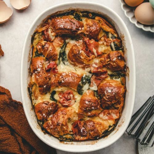 Overhead view of prosciutto and spinach strata casserole dish. The dish is surrounded by fresh eggs, egg shells, a hand mixer, various plates and a bowl cloth.