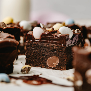 Close up view of a frosted brownie slice topped with mini eggs. Slice is surrounded by other pieces of brownie.
