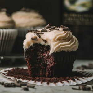 Close-up view of an Irish coffee cupcake. Cupcake is made of a espresso chocolate cake base, topped with an Irish liqueur buttercream frosting and chocolate shavings. The cupcake has a large bite taken from it, and is resting on an open cupcake liner. Behind it are two more cupcakes on the left and a bottle of Baileys on the right.