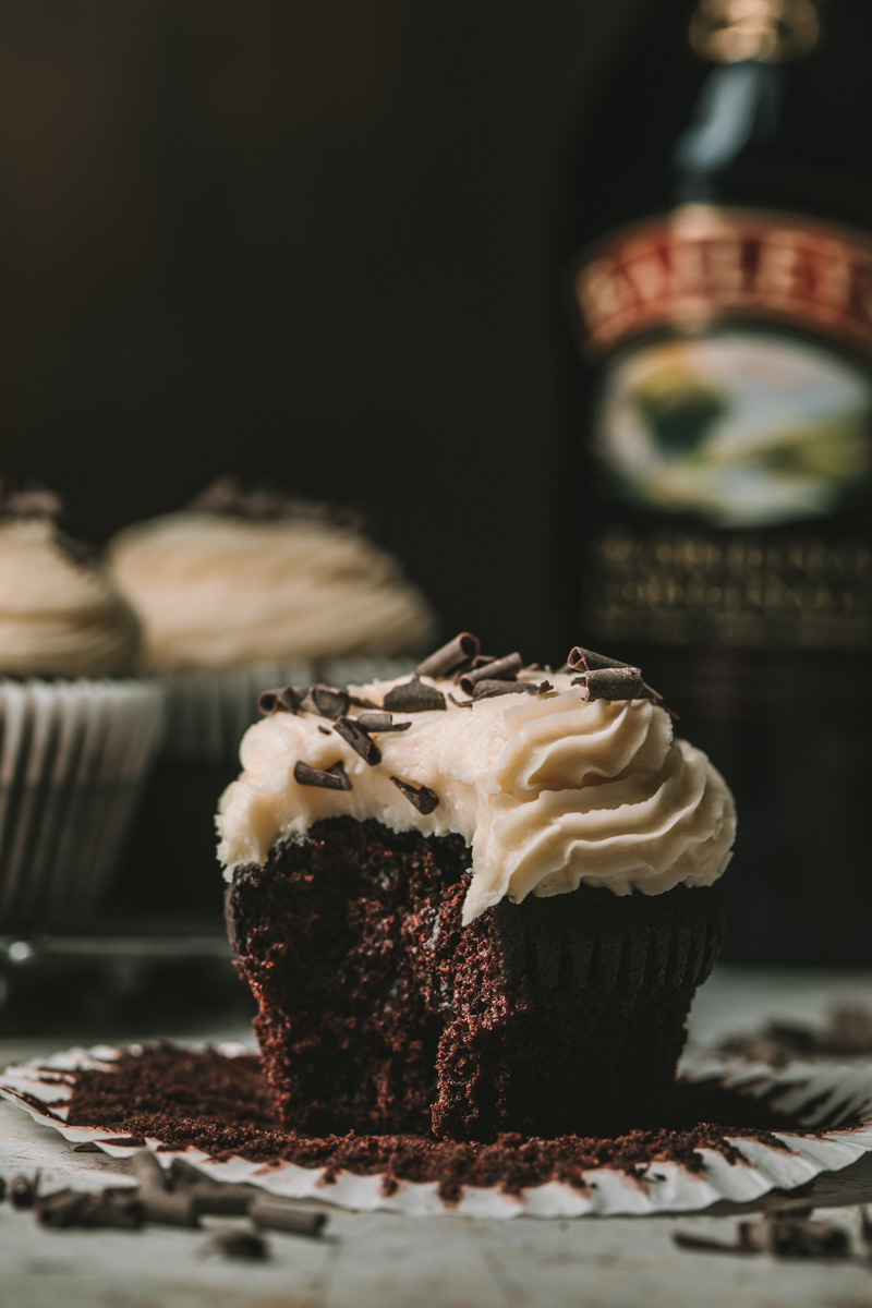 Close-up view of an Irish coffee cupcake. Cupcake is made of a espresso chocolate cake base, topped with an Irish liqueur buttercream frosting and chocolate shavings. The cupcake has a large bite taken from it, and is resting on an open cupcake liner. Behind it are two more cupcakes on the left and a bottle of Baileys on the right.