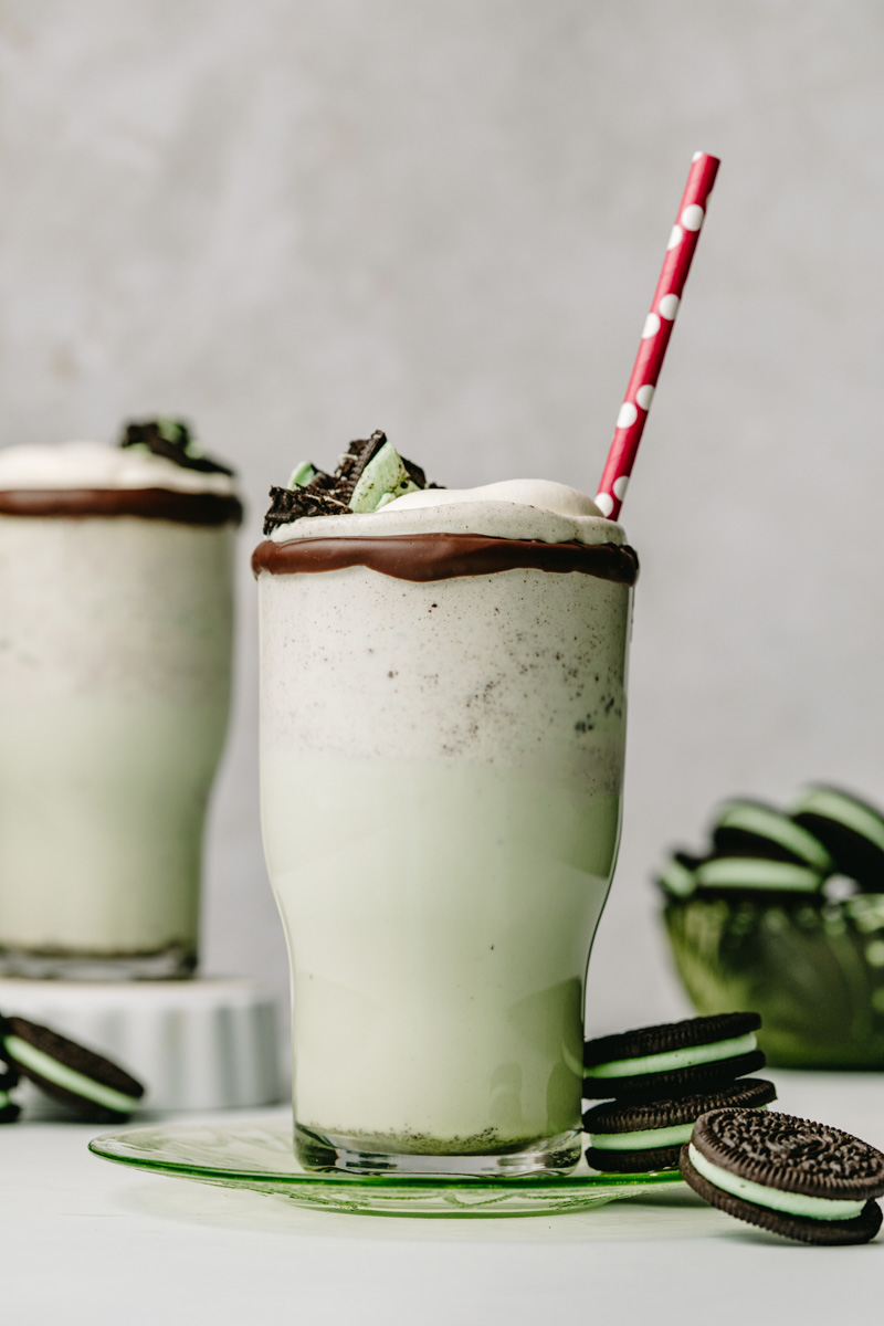 View of two oreo mint chocolate chip milkshakes. Both are garnished with whipped cream and oreo cookie pieces. The one closest to the viewer is resting on a green plate, and surrounded by oreo cookies.