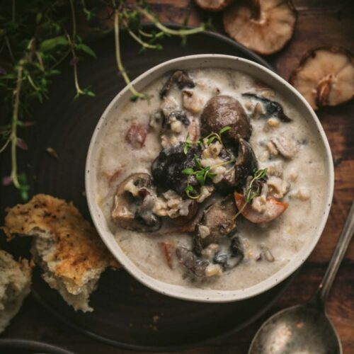 Overhead view of a bowl of vegetarian creamy mushroom and rice soup. The bowl is resting on a rustic wood surface, and is surrounded by fresh mushrooms, fresh thyme, a soup spoon and crusty bread.