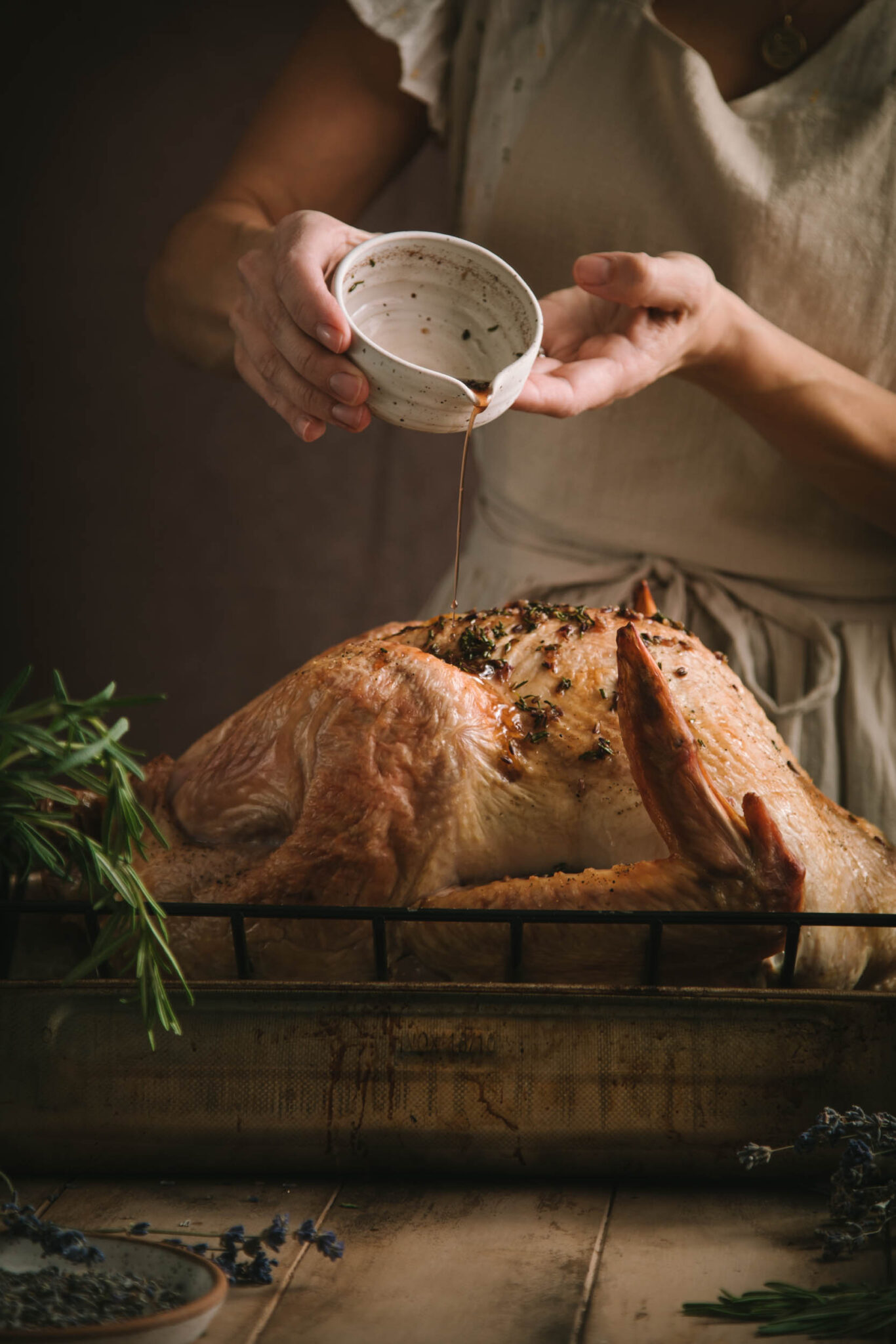 Woman in a beige linen apron pouring a fresh herb mixture onto a full turkey.