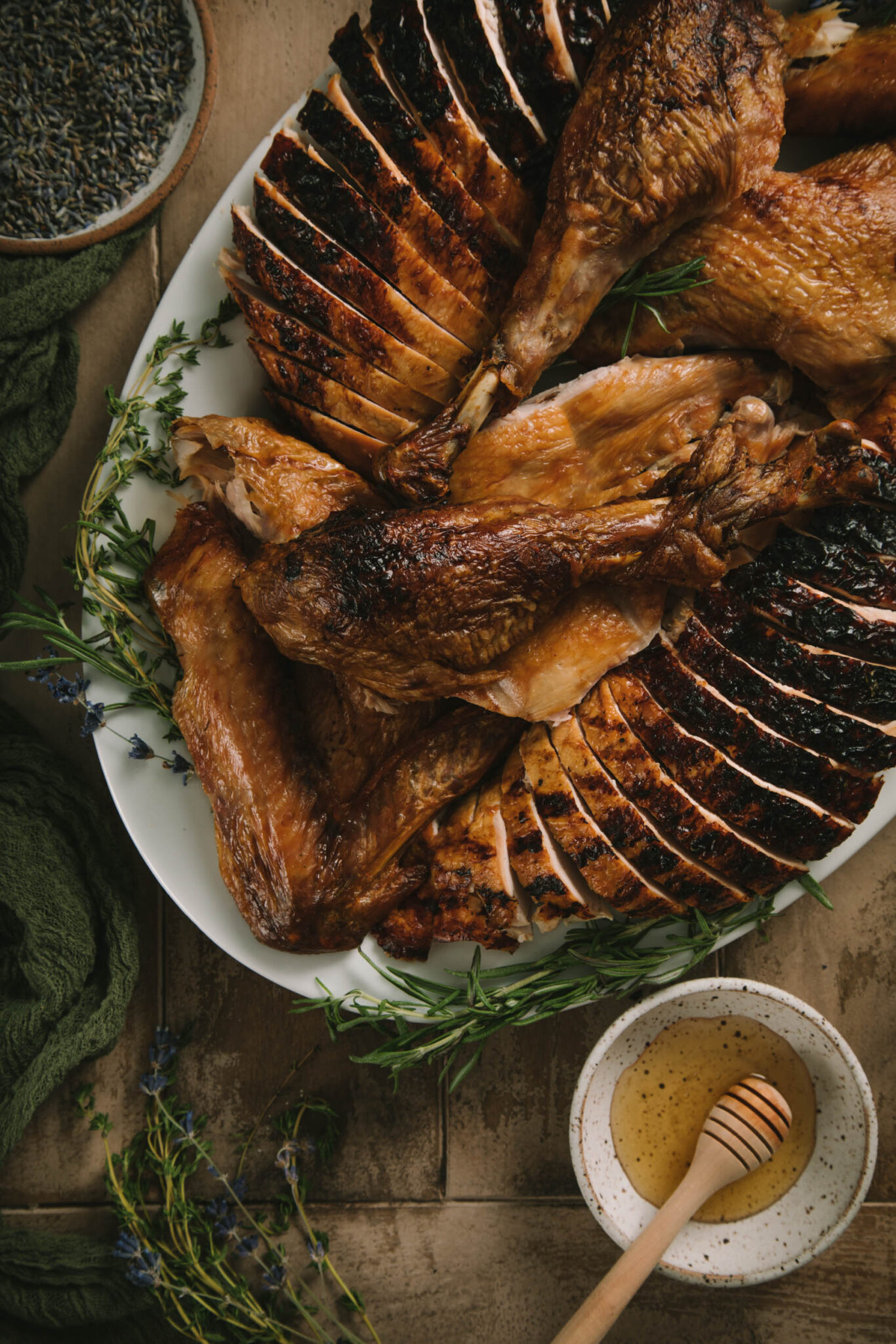 Overhead view of fully carved turkey on a rustic wood surface. Turkey is on a while serving plate. A small bowl of honey is to the right, and a dark green linen to the left.