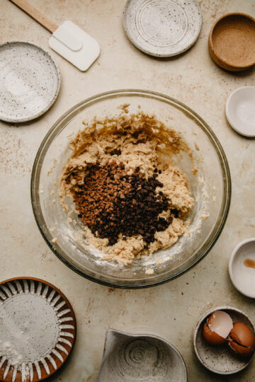 Overhead view of a large glass mixing bowl filled with cookie dough. Surrounding the bowl are various other small bowls and plates filled with ingredients. Over the batter are the mini chocolate chips and Skor bits.