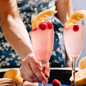 Tray of lemon raspberry mimosas, garnished with fresh berries and lemon rind, a hand reaching to grab a glass.