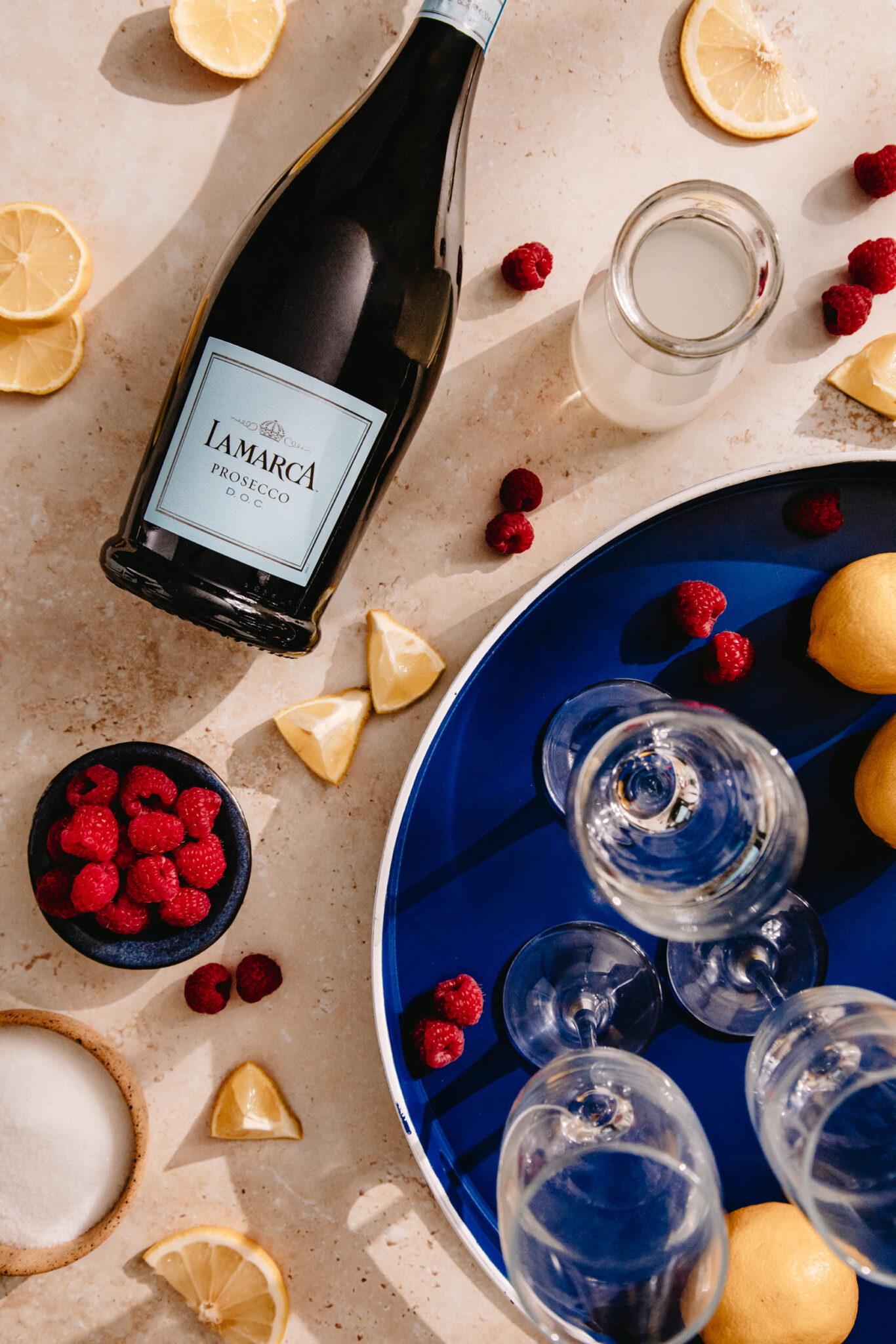 Overhead view of a blue tray with empty champagne glasses, a bottle of sparkling wine, and slices of lemon and fresh raspberries.