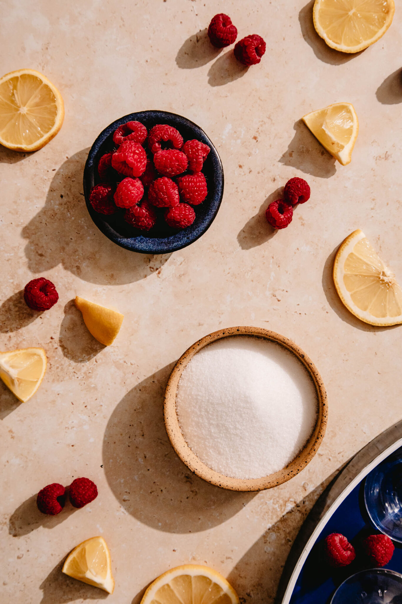 Overhead view of a bowl of granulated sugar and a bowl of fresh raspberries, surrounded by lemon slices.