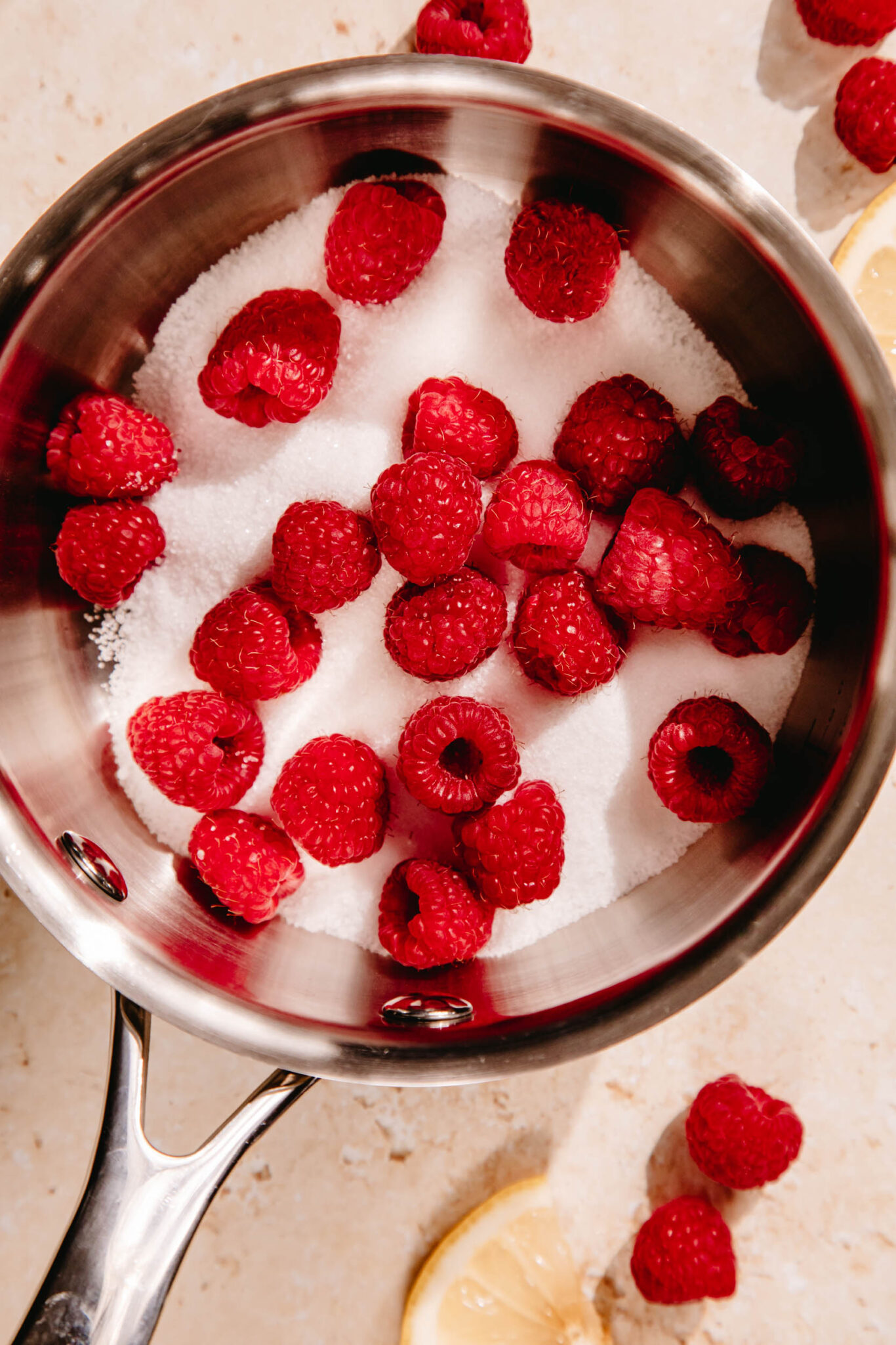 A small sauce pan filled with granulated sugar and fresh berries, on a beige backdrop.