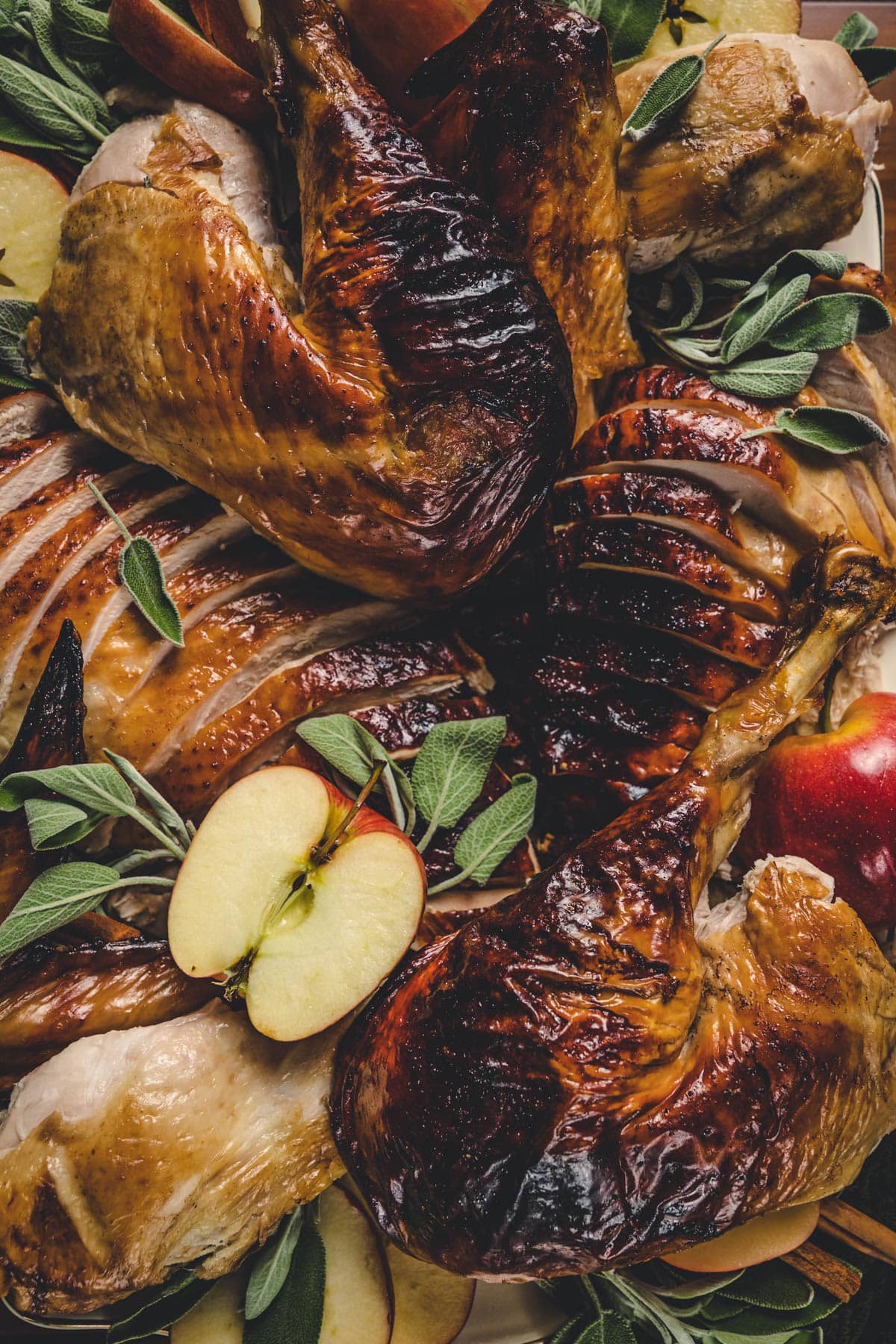 Apple honey glazed turkey, carved and placed onto a white serving platter with gold rim. Turkey is surrounded by cinnamon sticks, apples, and sage leaves.