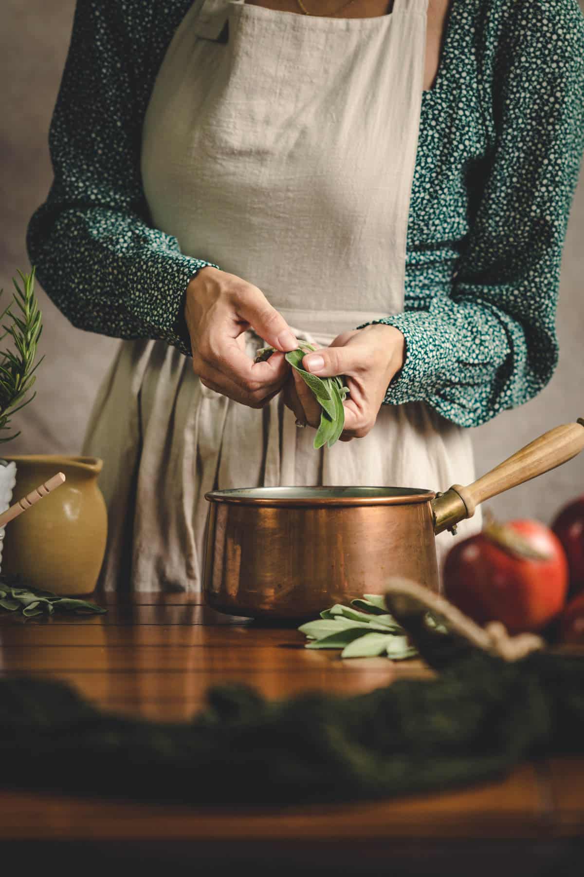 Woman in a green floral shirt and beige apron, pulling sage leaves into a brass saucepan.