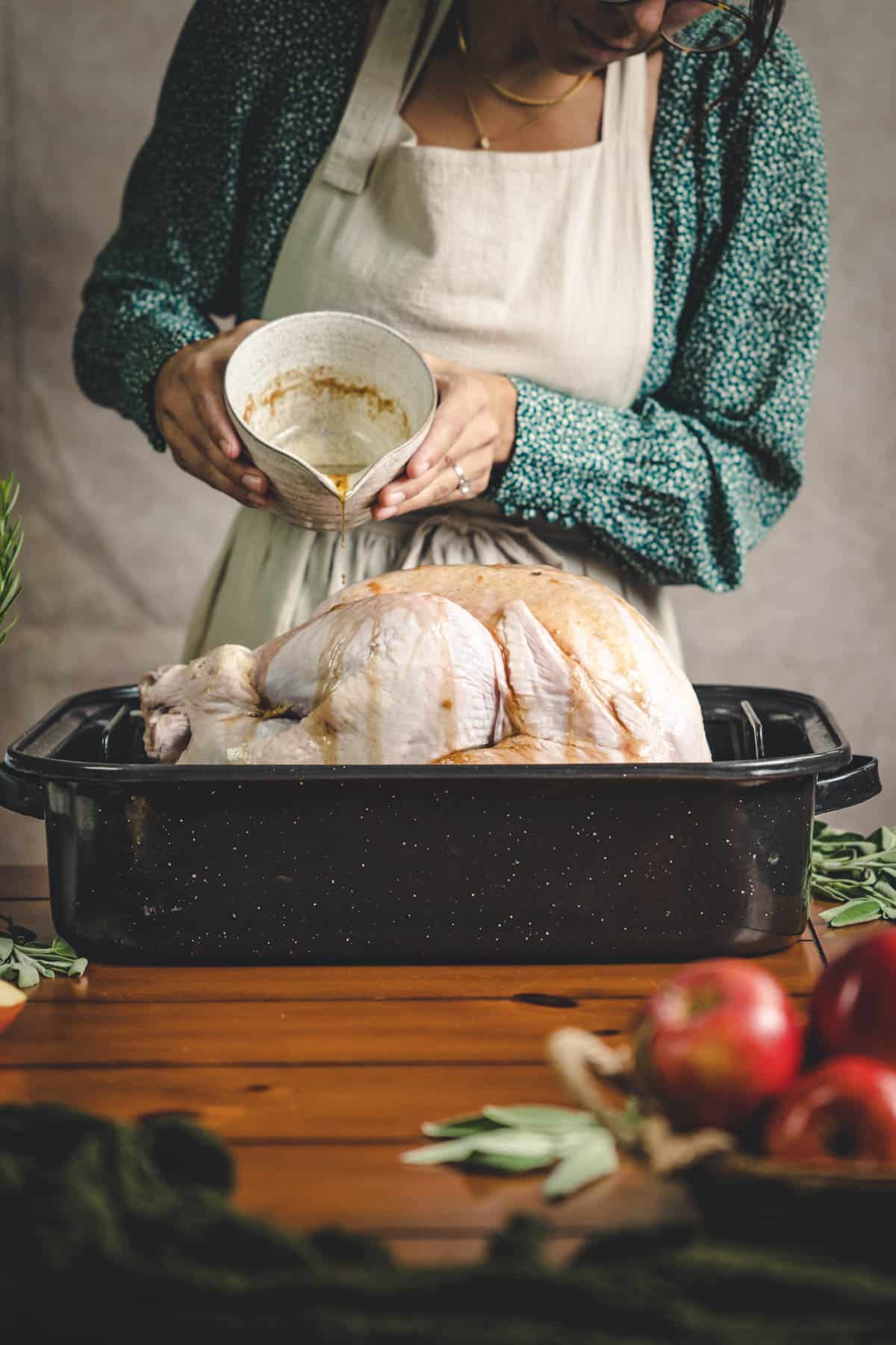 Woman in a green floral top and beige apron, pouring apple honey glaze onto a raw turkey.