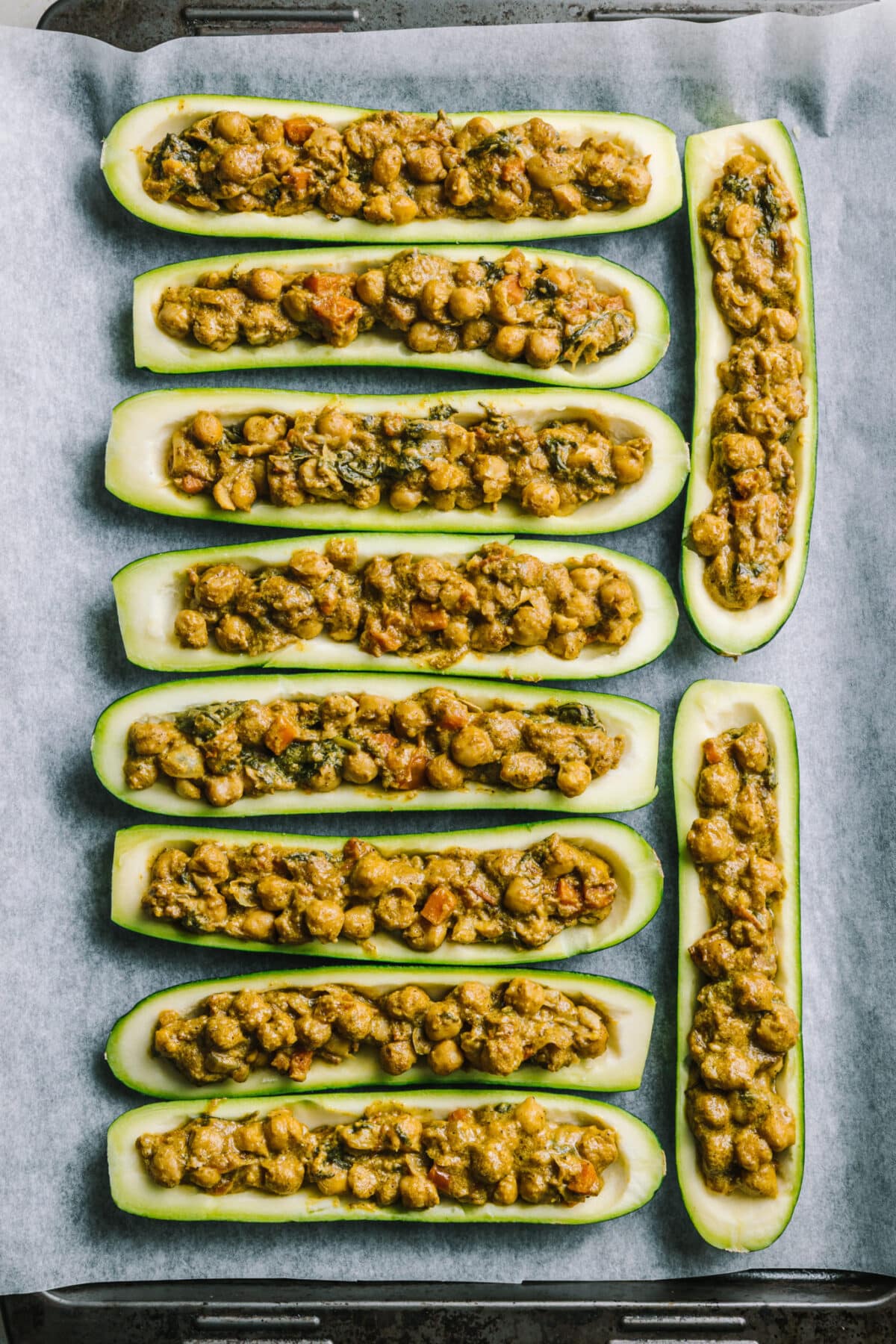 Baking pan lined with parchment paper, filled with uncooked chickpea curry stuffed zucchini boats.