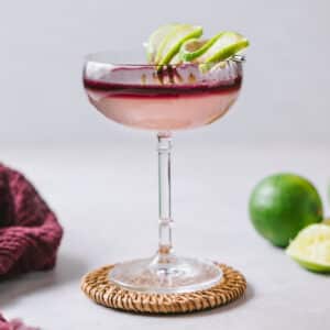 Front view of a tall coupe filled with a Devil's Margarita, surrounded by burgundy linen and limes.