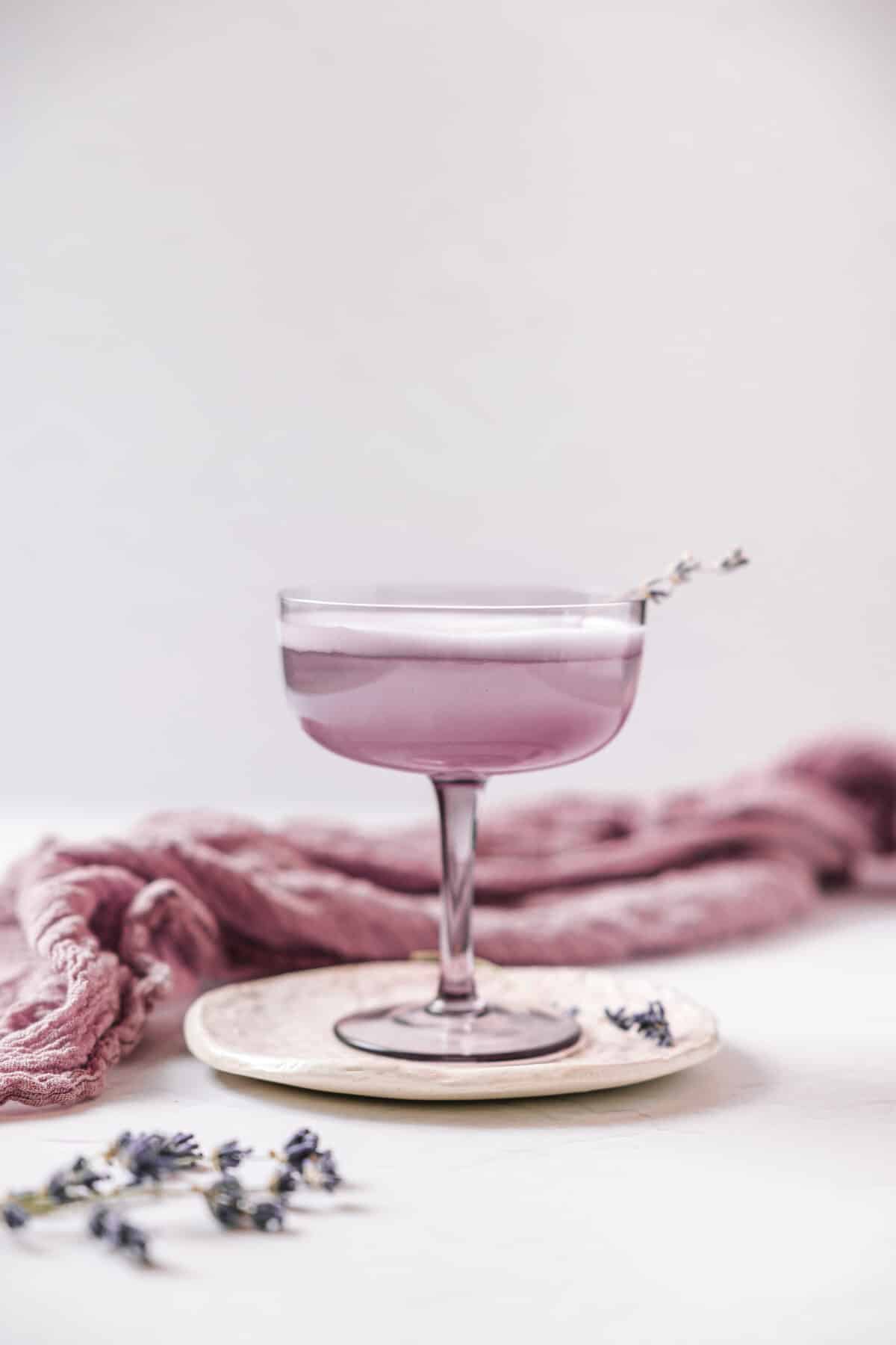 View of a lavender gin fizz, served in a purple coupe glass over a pretty floral print coaster. In the background is a lavender coloured linen. Lavender sprigs are in the left corner.
