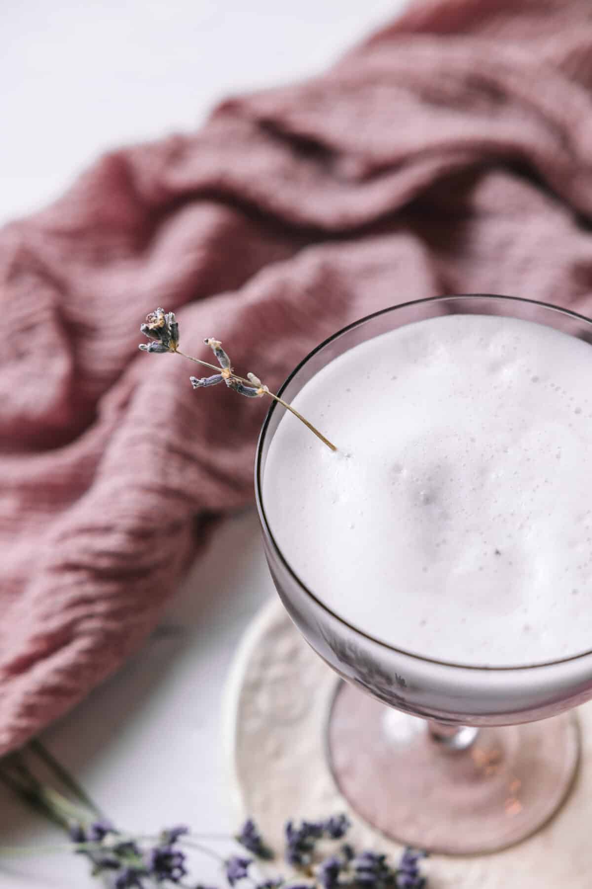 Close-up view of a lavender gin fizz, served in a purple coupe glass over a pretty floral print coaster. In the background is a lavender coloured linen. Dried lavender springs are found around the cocktail as well as used for garnishing.