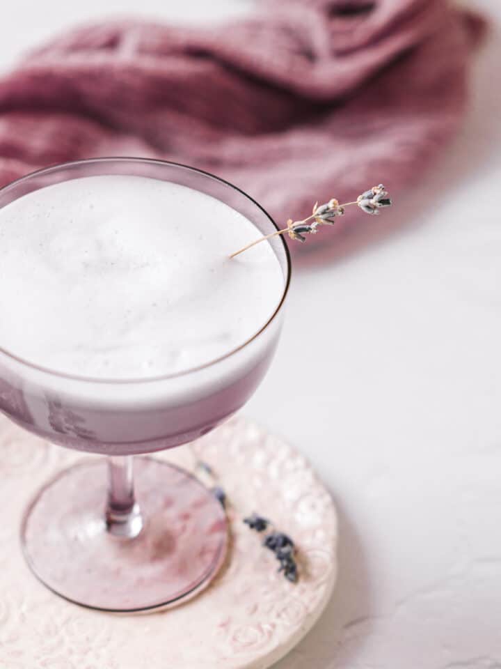 Angled view of a lavender gin fizz, served in a purple coupe glass over a pretty floral print coaster. In the background is a lavender coloured linen.