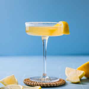 Direct view of a Sidecar cocktail in a martini glass, placed on a wicker coaster. The backdrop is blue, with sliced lemons surrounding the glass.